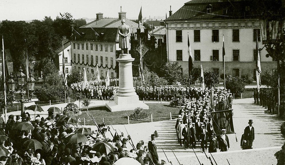 Studenter med fanor i procession genom universitetsparken mot universitetshuset förbi statyn över Erik Gustaf Geijer. I bakgrunden det Ekermanska huset och Gustavianum.