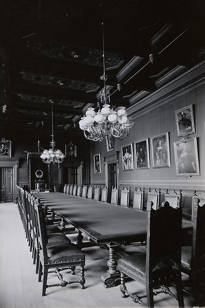 Black and white photo of the board's meeting room with long table, solid wooden chairs, chandeliers hanging from the ceiling and photos of kings on the walls