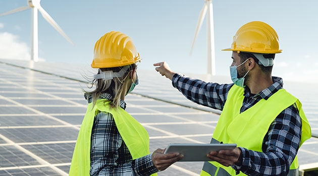 Two people in yellow vests and hard hats gaze over large solar panels.