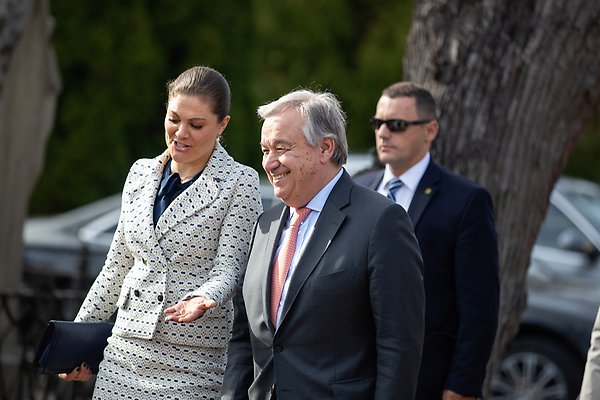 Crown Princess Victoria and UN Secretary-General António Guterres walk in a park and chat gleefully. Behind them is a costumed guard in sunglasses and some cars.