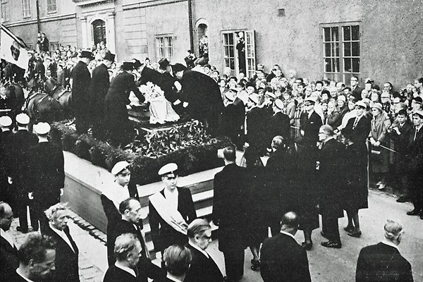 Men in black clothes and top hats place Dag Hammarskjöld's coffin on a raised stand with stairs. Students in student caps stand aroud it, a little further away crowd beyond a picket fence is watching the ceremony.