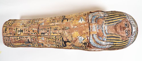 Hela sarkofagen, liggande på ett vitt bord. Den är i lera och målad i olika färger, ett tydligt ansikte med stora ögon längst upp. 