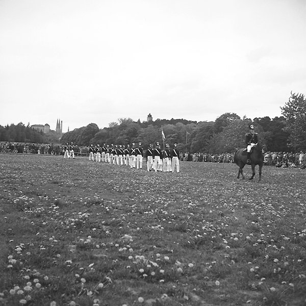Black and white photo of soldiers in formal dress marching in line and an officer sitting on a horse in front of them