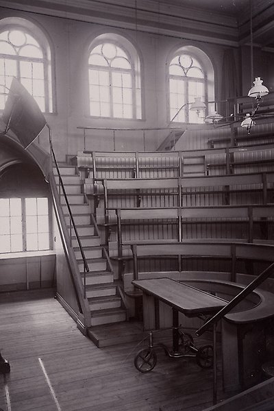 Black and white photo of lecture hall with tiered meadows forming a semi-circle around a table, a staircase leading to the rows and several windows at the back