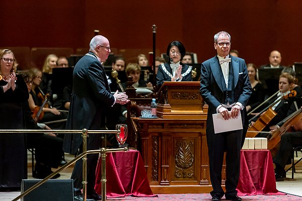 Kenneth Wärnmark stands with diploma in hand on the stage in the university hall. Next to him stands principal Anders Hagfeldt and applauds. Behind them, the deputy vice-chancellor Coco Norén in the lectern. Orchestra in the background.