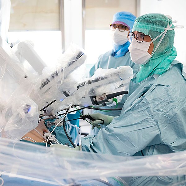 Två personer i heltäckande skyddskläder och en inplastad robot i en operationssal.