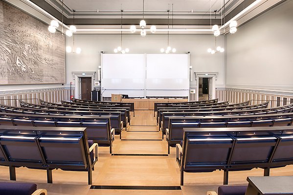 Hall IX seen from the aisle in the row at the back. A staircase runs along the central aisle. Behind the board are two whiteboards and two projection screens. On each side of the lectern there are two doors which are also emergency exits.