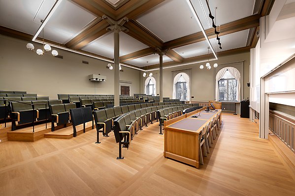 Hall IV seen from the side. At the front is the lectern with a table, chairs and a pulpit. Behind the whiteboards and projection screens. On it are microphones and a screen where you manage sound and the projector. The rows of benches form a slightly curved arch. The two front rows are at ground level and on the far right there are three wheelchair spaces. The rows behind slope slightly upwards. They are divided into three sections with two aisles with stairs.